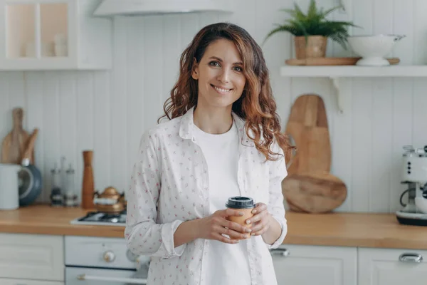 Stock image Relaxed smiling hispanic woman in cozy kitchen holds coffee, looks at camera. Pleased homeowner enjoys domestic routine at home