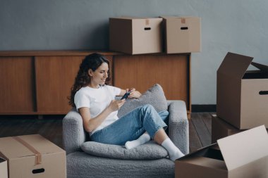 Taşınma günü: İspanyol kız karton kutuların arasında oturuyor, akıllı telefona bakıyor, internetten mobilya alışverişi yapıyor veya taşınma hizmetleri düzenliyor.