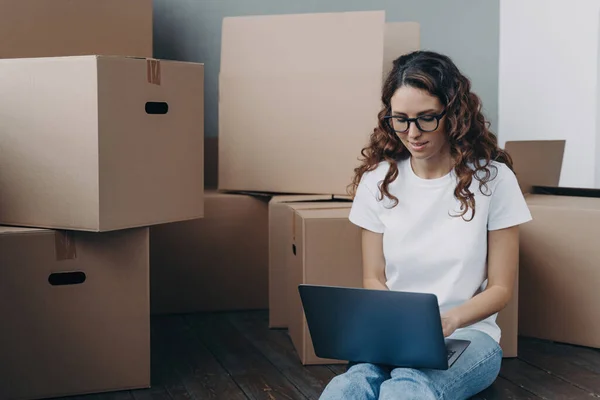 Focused woman in glasses reviews moving arrangements online, sitting amidst cardboard boxes, searching for a reliable mover.