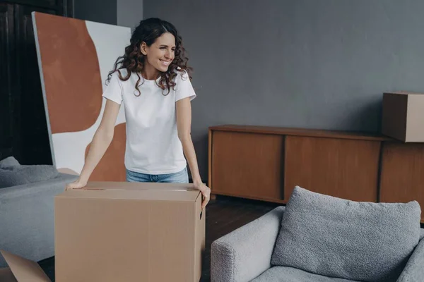 Happy young Spanish girl in new home apartment with a cardboard box. Pleased woman prepares for house renovation or relocation, dreaming of modern interior design.