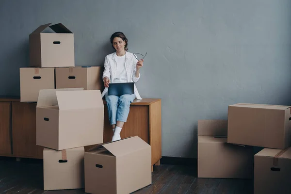 Female with laptop surrounded by cardboard boxes, searching for a moving company. Woman selects mover for relocation. Transportation services concept.