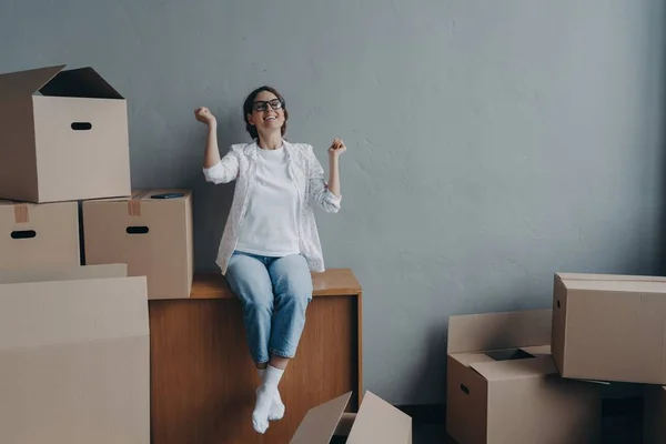Excited Spanish girl celebrating relocation to new home. Joyful woman sitting with cardboard boxes, happy with moving day. Real estate purchase concept.