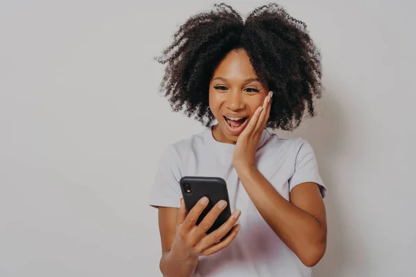 stock image Excited mixed race ethnicity millennial girl isolated on white studio background feeling delightfully winning on cellphone app, overjoyed happy african young woman gets great news on smartphone