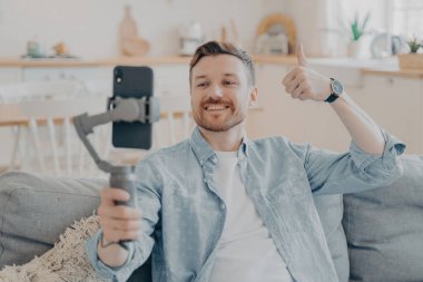 Videodaki tatmin olmuş genç adam, telefonu elinde tutuyor, başparmağını kaldırıyor, kanepeye oturuyor.