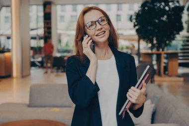 Kendine güveni tam, birden fazla iş yapan, dizüstü bilgisayarı ve defteri olan, resmi giyinmiş ve gözlük takmış bir kadın girişimci müşteriyle hoş bir telefon görüşmesi yapıyor..