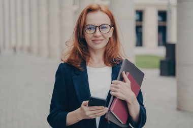 Gözlük takan, akıllı telefon ve dizüstü bilgisayar tutan, kendine güvenen kızıl saçlı kadın bir iş danışmanı, dışarıda gülümseyerek poz veriyor..