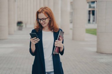 Verimli bir günün ardından cep telefonu, dizüstü bilgisayar ve dizüstü bilgisayar kullanarak açık havada şık giyinen kızıl saçlı olumlu bir iş kadını..