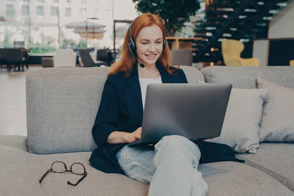 コーヒーショップでの笑顔の赤毛のフリーランスは オンライン言語のレッスンを提供し 無線ヘッドセットを着て ノートパソコンのビデオ通話でクライアントと通信します — ストック写真