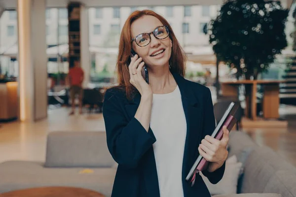 自信を持って生姜の女性起業家マルチタスク ノートパソコンやノートブックを保持 クライアントとの快適な電話の会話を持っています 正式な服装や眼鏡を身に着けています — ストック写真