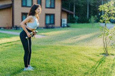 Üstte sportif bir kadın, dambıllarla spor yapıyor, pazılarla çalışıyor, özel bir evin yanında açık havada çalışıyor. Etkin yaşam biçimi.