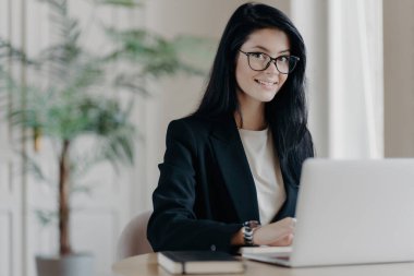 Gülümseyen kadın sekreter modern bir ofis masasında dizüstü bilgisayarda çalışıyor, proje yürütüyor ya da araştırma yapıyor. Konforlu uzak görev ayarları.