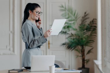 Gri kostümlü mutlu kadın yönetici kağıtları kontrol eder, göz giysisi giyer, araştırmaya odaklanır, iş yerinde telefon görüşmesi yapar..