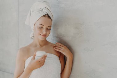 Tatmin olmuş kadın yüz kremi sürüyor, havluya sarılmış, gri arka plan yakınlarında. Sauna sabahı rutini.