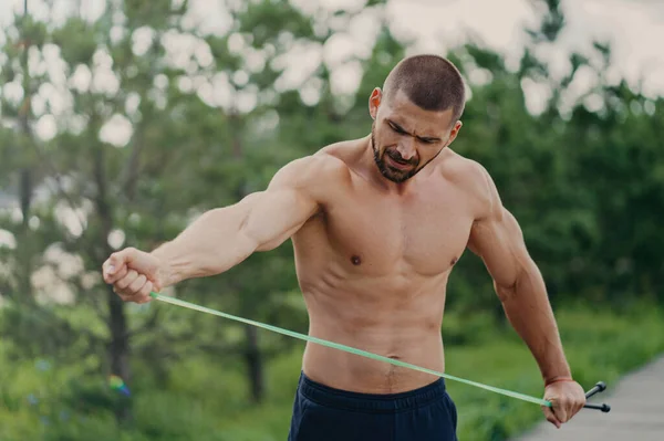 stock image Handsome man with naked torso poses at park, stretches skipping rope, ready for outdoor workout. Street workout concept.