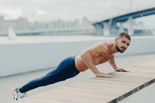 スポーツウェアの板の筋肉の男は 屋外でプッシュアップを行い 強い腕のための決意を示しています — ストック写真