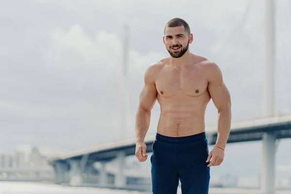 stock image Confident bearded man with naked torso poses by river bridge, leads healthy lifestyle, has muscular body.