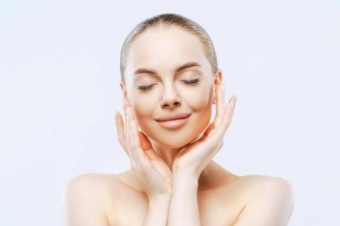 Güzel bir kızın yakın plan portresi, gözleri kapalı, şefkatle dokunan yüz, parlak ten rengi, sağlıklı ten, beyaz duvara karşı üstsüz. Tazelik, vücut bakımı.