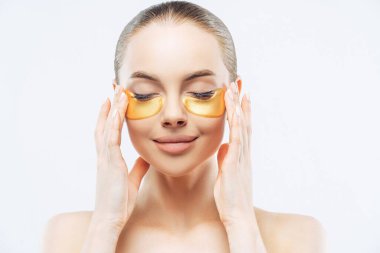 Gülümseyen Avrupalı kadın yaşlanmayı önleyici göz terapisi, altın bantlar, temiz cilt, beyaz arka planda üstsüz.