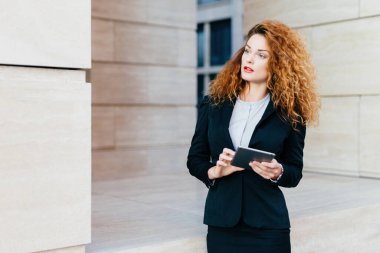 Kıvırcık saçlı girişimci resmi kıyafet planları yapıyor, iş günü tablet tutuyor, dalgın dalgın bakıyor. Touchpad ile proje geliştiriliyor.