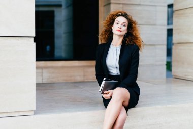 Kendine güvenen, dalgalı saçlı, kırmızı dudaklı, resmi giyinmiş, tablet tutan, bacak bacak üstüne atan, iş havası yayan bir kadın model..