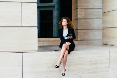 Düşünceli bayan girişimci, siyah resmi kıyafet, bacak bacak bacak üstüne atma, tablet tutma, gelecekteki ortaklıkları öngörme.