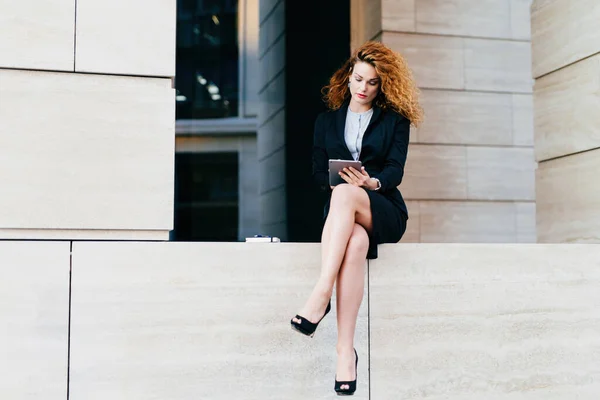 stock image Curly-haired model, elegant suit, high heels, slender legs. Uses tablet for communication. Freelancer works outdoors, looks serious.