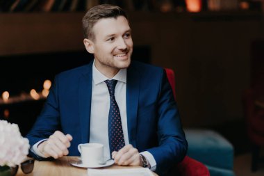 Kafede neşeli bir işadamı, kahve içiyor, mutlu, birini fark ediyor, hoş bir gülümseme, resmi kıyafetler.