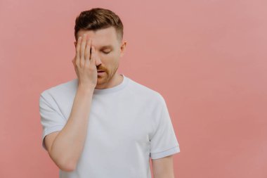 Beyaz tişörtlü yorgun bir adam yüz avuç içi yapar. Sıkılmış, pembe arka planda izole edilmiş hisseder.