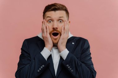 Şok olmuş kızıl saçlı adam, şaşkın ifade, inanılmaz haberler alıyor, çenesi düşük, pembe arka planda poz veriyor.