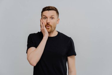 Siyah tişörtlü, şaşkın, şaşkın kızıl adam. Stüdyo görüntüleri, kameraya şaşkın bakışlar. Gri duvar arkaplanı. Duygular konsepti.