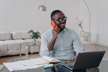 Mutlu Afrikalı Amerikalı iş adamı, iş görüşmesi yapıyor, ofis masasında dizüstü bilgisayarla çalışırken cep telefonuyla konuşmaktan hoşlanıyor..