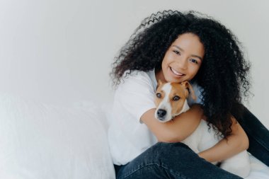 Afro 'su olan genç bir kadın, köpeğini kucaklar, ilgilenir, gülümser, günlük kıyafetler, beyaz arka plan, yatak, tanıtım alanı. 