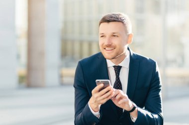 Delighted thoughtful elegant male marketing trader in formal black suit, uses modern cell phone for searching website in internet, types text message, looks pensively into distance. Bussiness concept