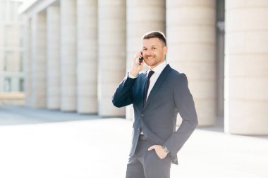 Horizontal shot of satisfied male with positive look solves banking problems while calls to operator via smart phone, wears elegant black suit, poses putdoor in urban setting. Communication concept