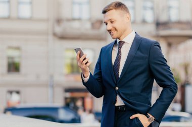 Horizontal shot of happy businessman keeps hand in pocket, wears formal suit and wristwatch, recieves notification on smart phone, finds out about high financial balance. Cheerful trader outdoor