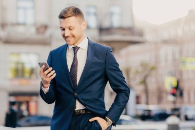 Young succesfful delighted happy businessman reads text message on modern cellular, wears elegant suit, holds hand in pocket, enjoys online communication. People, business, technology concept