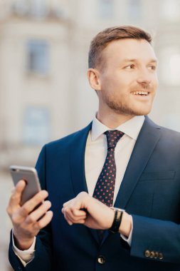 Attractive male comes on meeting with client or partner who is late, looks at watch and holds smart phone, looks aside, hopes to see someone into distance, dressed in formal suit, stands outdoor