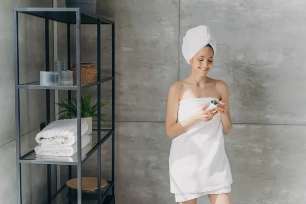 ヨーロッパの女性は瓶からクリームを適用します 入浴後のタオルで魅力的な女の子 自宅で幸せな女性のシャワー スキンケアのコンセプト — ストック写真