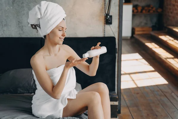 タオルを着た女性はベッドの上に座っている 女の子は ボトルの後風呂からボディ保湿剤を適用します 朝のシャワー 自宅で毎日のスキンケア — ストック写真