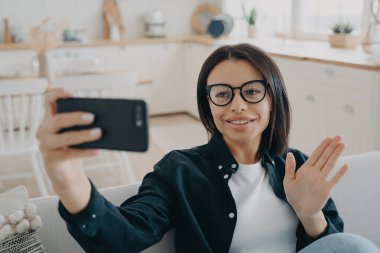 Gülümseyen kadın video çağrısına cevap veriyor, el sallıyor, kanepeye oturuyor. Genç blogcu internette sohbet ediyor, takipçileri karşılıyor.