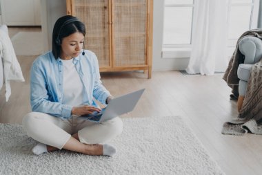 Odaklanmış serbest çalışan dizüstü bilgisayarla çalışıyor, evde yerde oturuyor. Kablosuz kulaklık takan ciddi bir kadın eğitici web sitelerine katılıyor, internetten okuyor. E-öğrenme, uzak meslek.