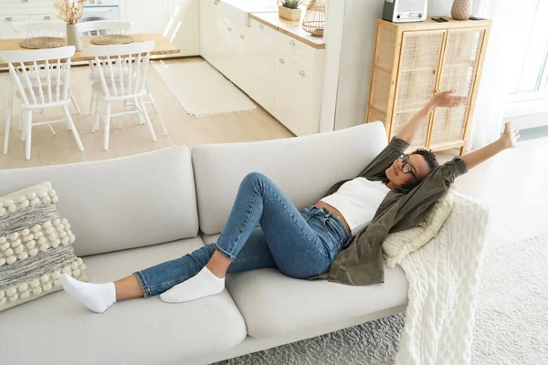 stock image Pleased African American girl stretching on a comfortable sofa at home, waking up after an afternoon sleep. Resting in a cozy living room, feeling lazy on a sunny weekend.