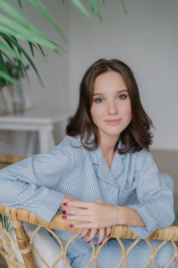 Pleasant young European woman with dark hair, striped jacket, red manicure, charmingly looks at camera, posing on wicker chair indoors.