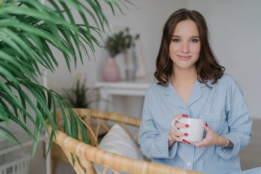 Image of relaxed woman in night costume, holding a cup of coffee or tea, enjoying morning in a spacious room with green plants. Spare time and leisure concept.