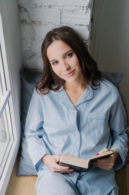 Beautiful relaxed woman in nightclothes, carefree expression, reading a book on a window sill in the morning, enjoying spare time and her hobby. Vertical shot of a teenage girl with literature.