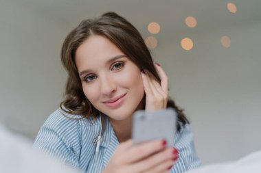 Caucasian woman in bed, using modern smartphone with earphones to watch videos and listen to audio on social networks. Technology and entertainment concept.