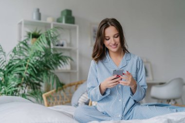 Happy dark-haired woman in casual attire, enjoying rest time in cozy bedroom. Listening to music with modern devices, domestic interior in background. Lifestyle concept.