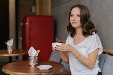 Carefree woman in a white t-shirt, enjoying aromatic coffee at a restaurant. Happy expression, thoughtful gaze into the distance while spending free time at a round wooden table.