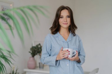 Young European woman in nightclothes, holding a white mug of coffee or cappuccino. Weekend morning in the living room, enjoying a good rest and leisure time. People and lifestyle concept.