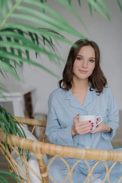 Adorable young woman in striped domestic costume, with a charming look, enjoys her day off, sipping a hot beverage in a wicker chair. People, lifestyle, and recreation time concept.